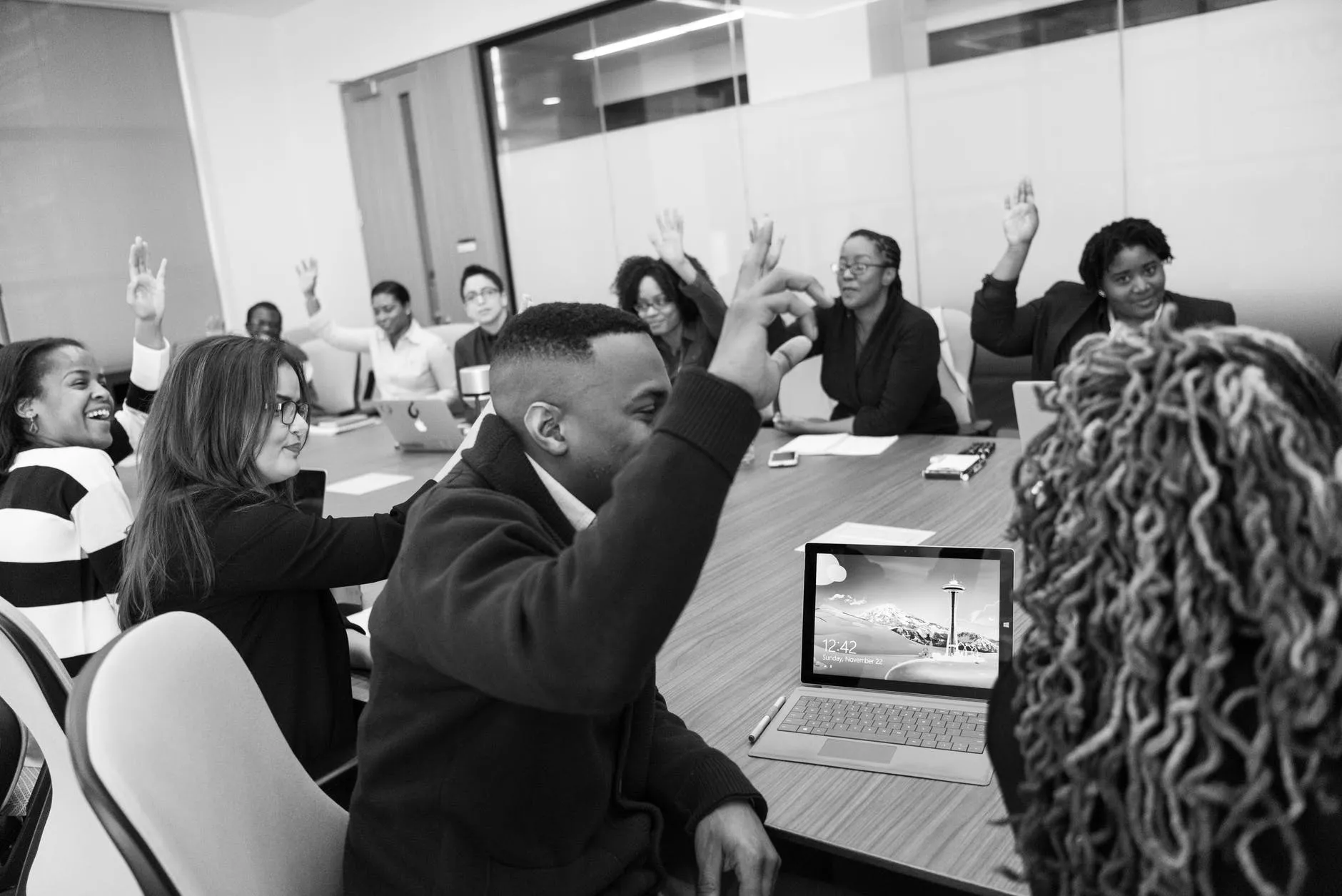 Black and white photo of people seated around a conference room with raised hands